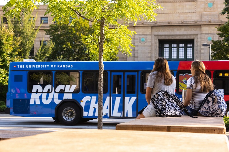 Bus reading "Rock Chalk" passes by two seated students outside Strong Hall